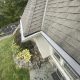 Downward view along a white gutter and shingle roof edge overlooking a stone patio and landscaped yard.